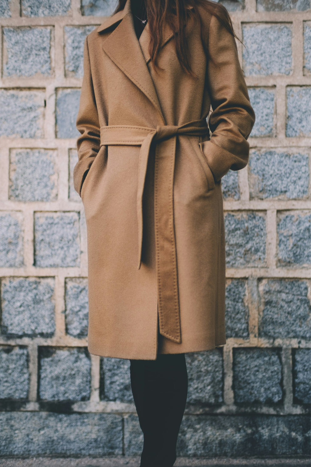Woman wearing a belted camel wool wrap coat in front of a stone wall, natural light