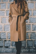 Woman wearing a belted camel wool wrap coat in front of a stone wall, natural light