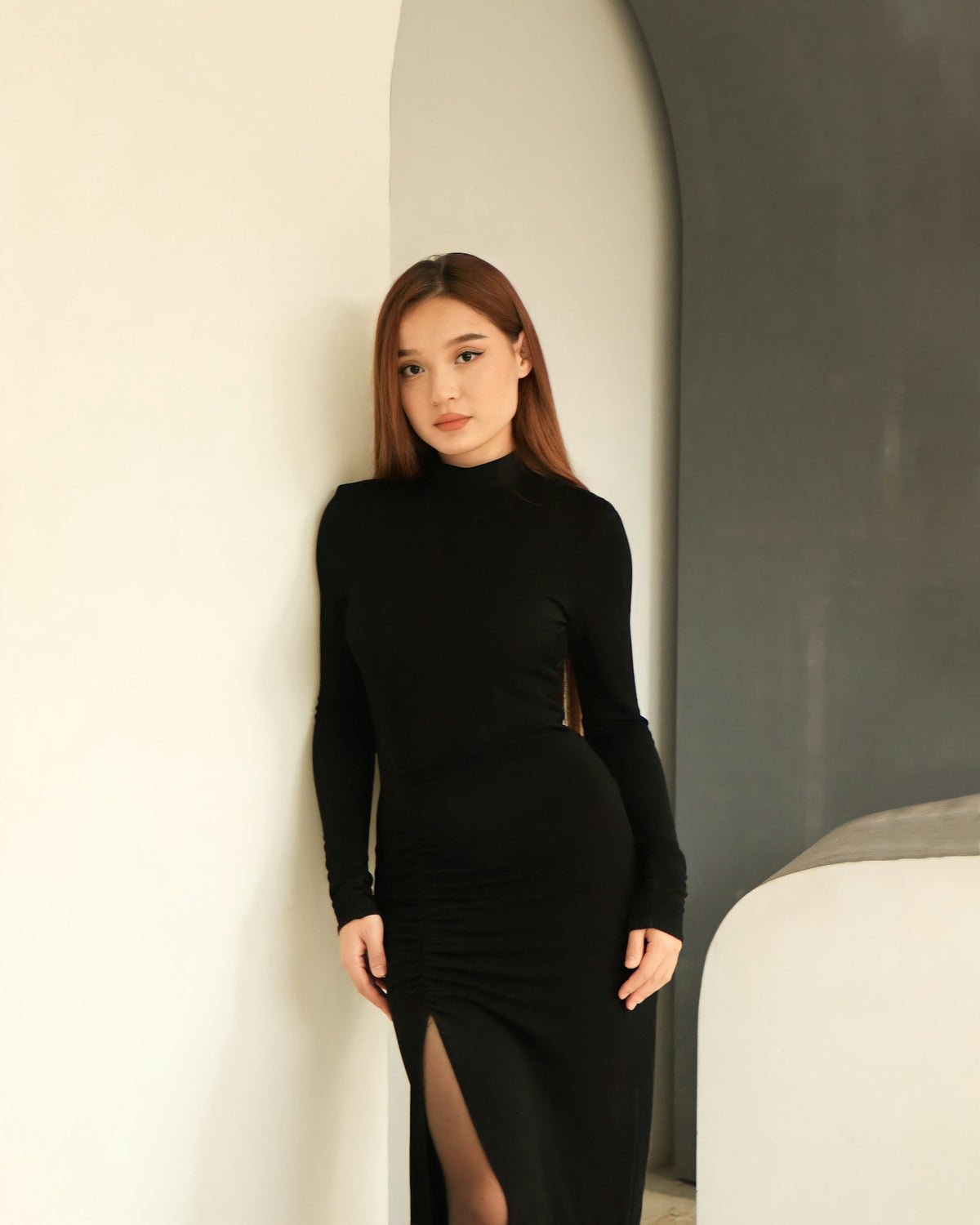 Woman in a long-sleeved black column dress leaning against a warm neutral wall